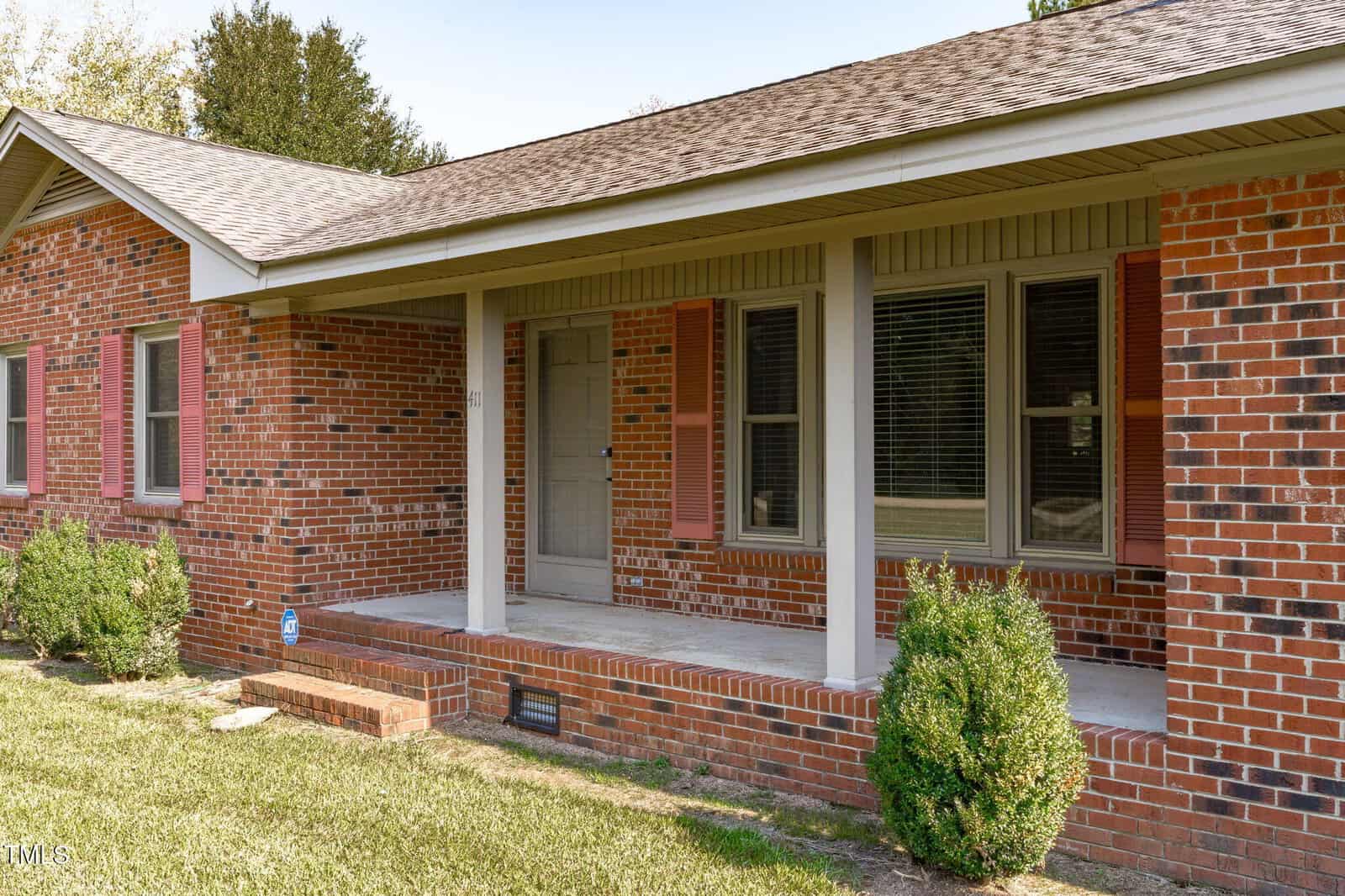 Single-story red brick house with a covered front porch, three shuttered windows, an off-center front door, small shrubs, and a neatly trimmed lawn. The roof has brown shingles and the porch has concrete flooring.