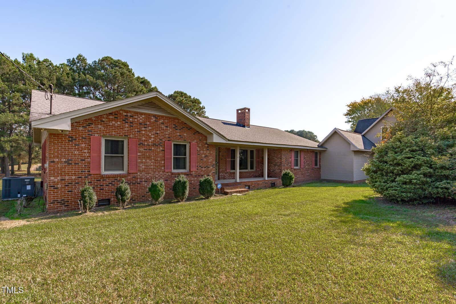 A single-story brick house with red shutters, a covered front porch, and a chimney. The front yard has trimmed grass, small bushes, and a few trees. There is a detached structure in the background.