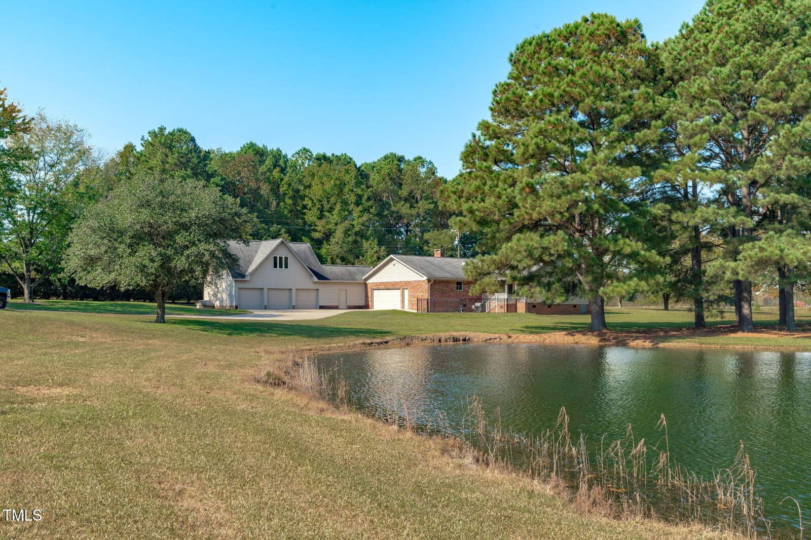 A house and detached garage sit among trees near a small pond, with a grassy yard in the foreground and a clear blue sky overhead.