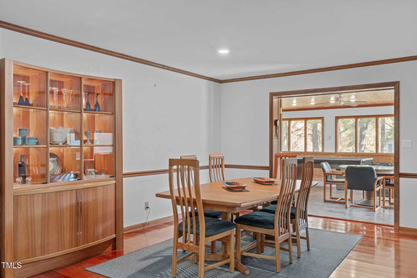 A dining room with a wooden table and six chairs sits on a gray rug. A built-in glass-front cabinet displays dishes. An adjacent room with additional seating and large windows is visible in the background.
