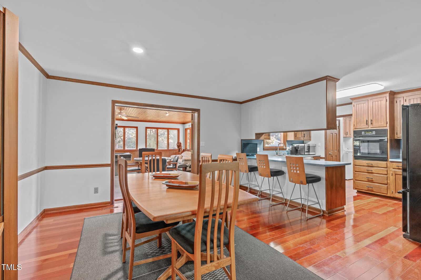 A dining area with a wooden table and chairs is adjacent to a kitchen with bar stools, wooden cabinets, and modern appliances. An adjoining room with large windows and a seating area is visible in the background.
