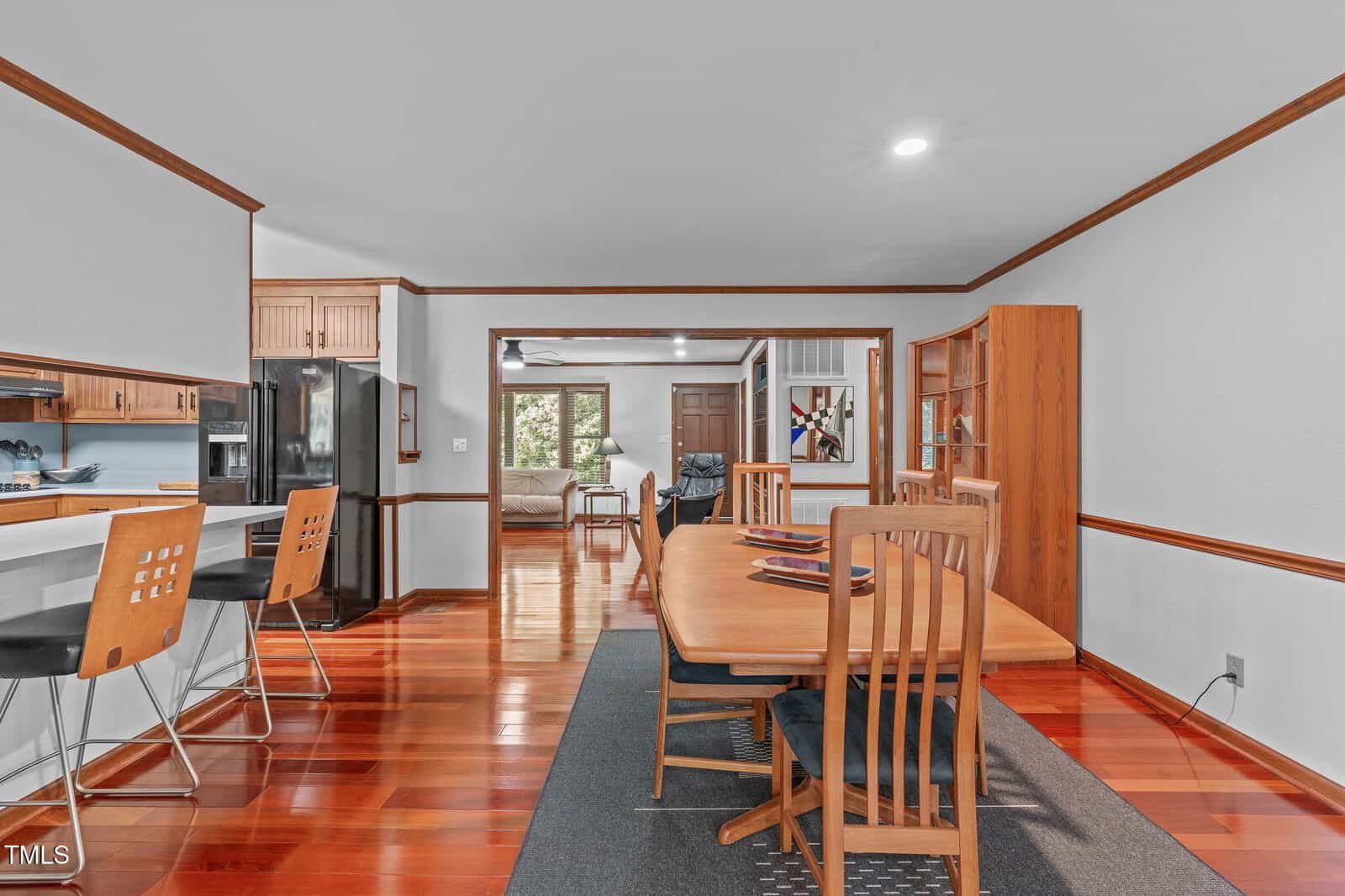 A dining area with a wooden table and chairs on a dark rug, adjacent to a kitchen with barstools and visible appliances. Beyond the dining area, a living room and additional seating are visible.