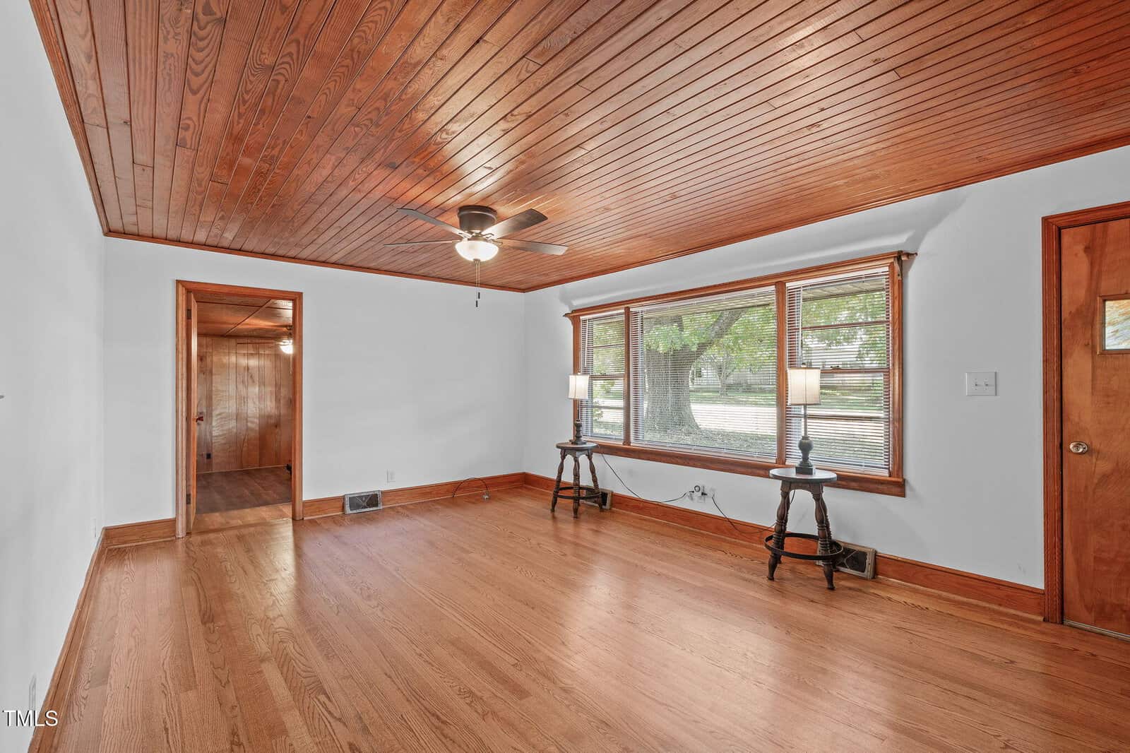 A living room with hardwood floors, wood-paneled ceiling, and white walls. Two windows with wood trim face outside, flanked by two lamps on side tables. A ceiling fan is centered on the ceiling.