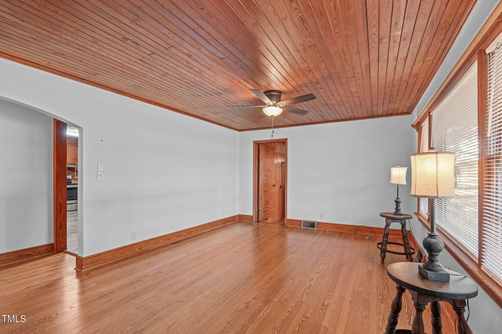 A spacious, empty living room with hardwood floors, wood-paneled ceiling, a ceiling fan, white walls, large windows with blinds, and two side tables with lamps near the windows.