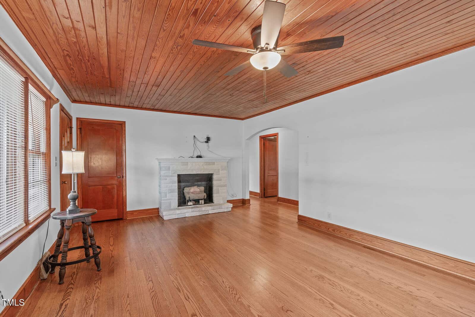 A living room with wood floors, a wood-paneled ceiling, a ceiling fan with a light, white walls, a white brick fireplace, a wooden side table with a lamp, and two windows with blinds.