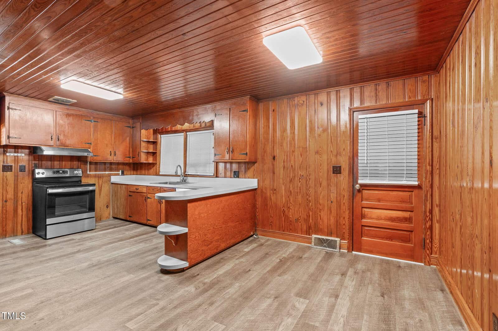 A kitchen with wood-paneled walls and ceiling, light wood flooring, stainless steel stove, white countertops, open shelves, a window above the sink, and a wooden door with blinds.