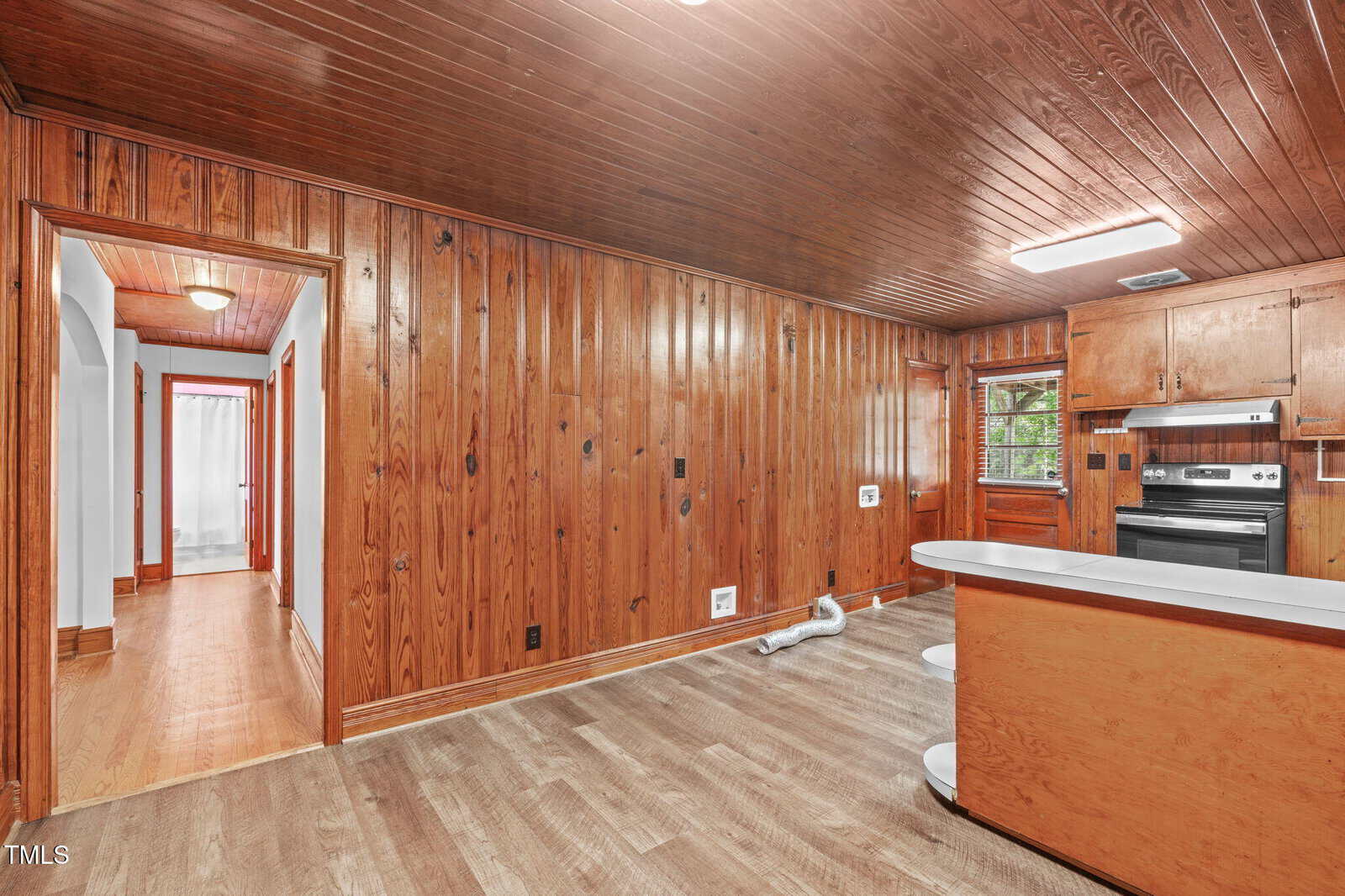 A room with wood-paneled walls and ceiling, wood-style flooring, and a kitchen area with wooden cabinets and black appliances. A hallway with wood floors and white walls is visible on the left.