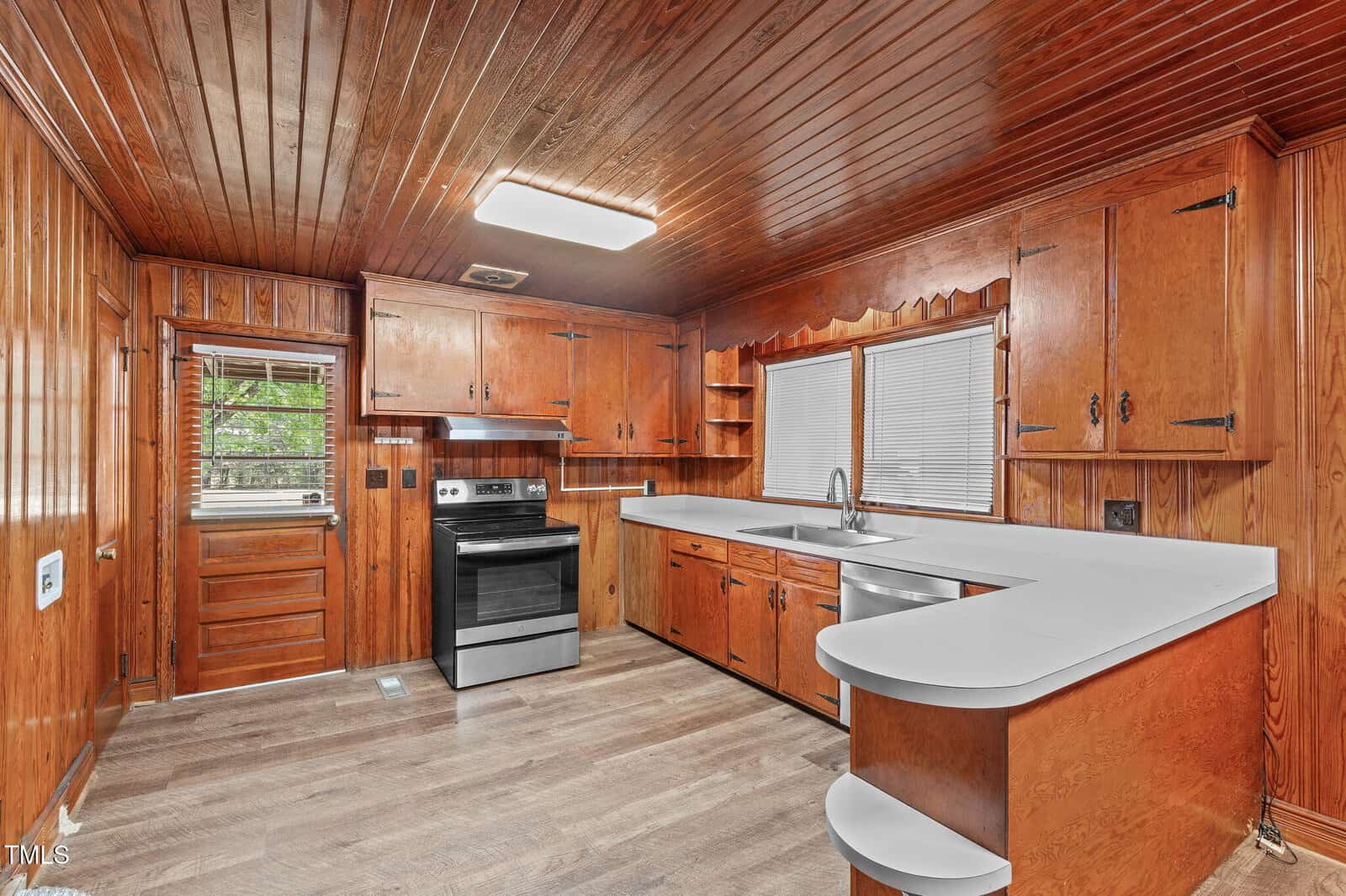 A kitchen with wood-paneled walls and ceiling, white countertops, a stainless steel electric stove, double sink under two windows, and a small attached breakfast bar. There is a door and window on the left side.