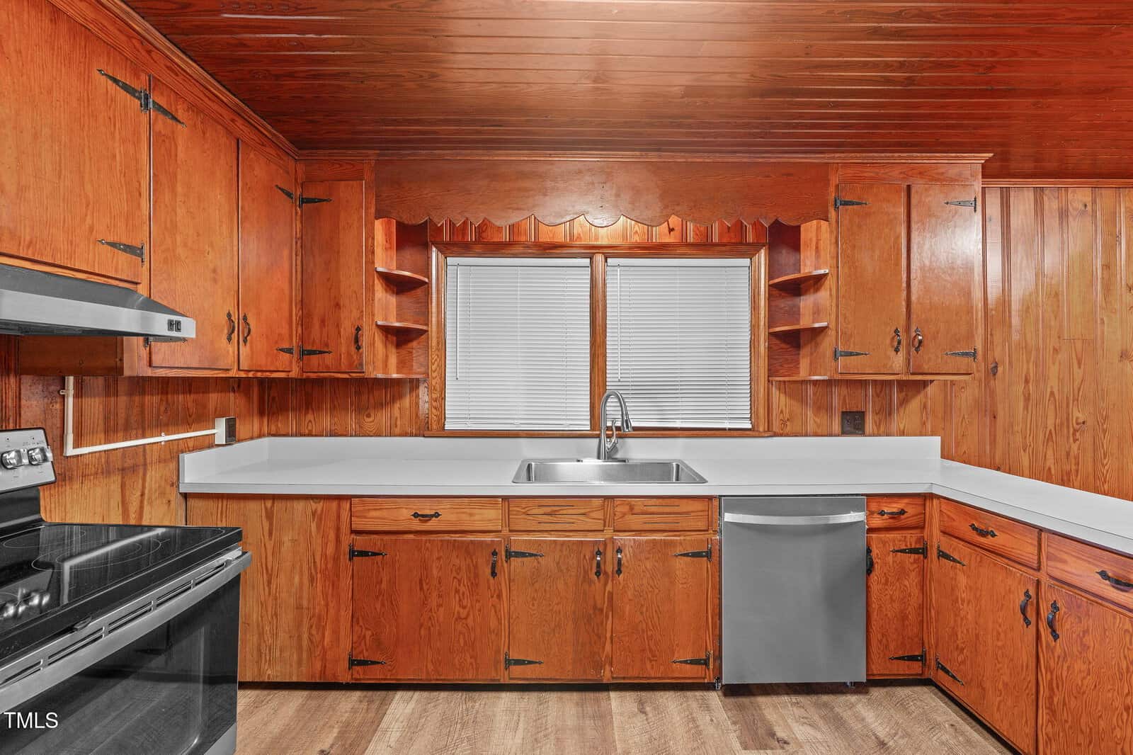 A kitchen with wood-paneled cabinets, white countertops, a stainless steel stove and dishwasher, a double sink under a window with blinds, and light wood flooring.