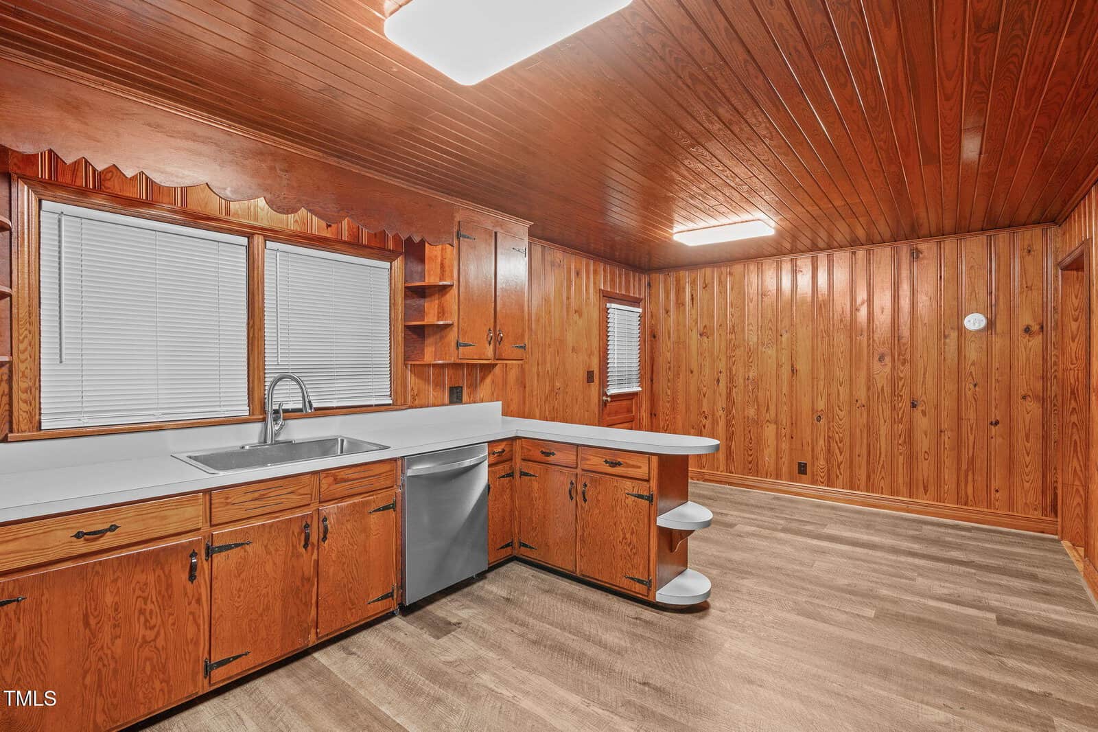 A kitchen with wood-paneled walls and ceiling, white countertops, wooden cabinets, a stainless steel sink, dishwasher, window blinds, and vinyl flooring. The space is well-lit with ceiling lights.