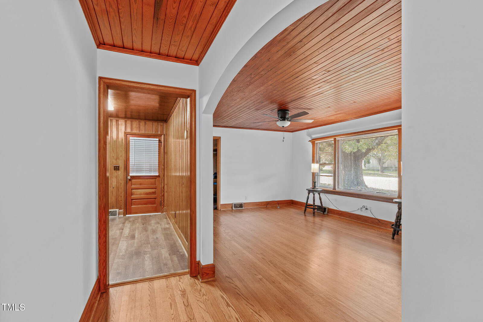 A spacious room with wood floors and ceiling, large front window, white walls, and a hallway with wood paneling and a closed door at the end. Ceiling fan and lamp visible in the main area.