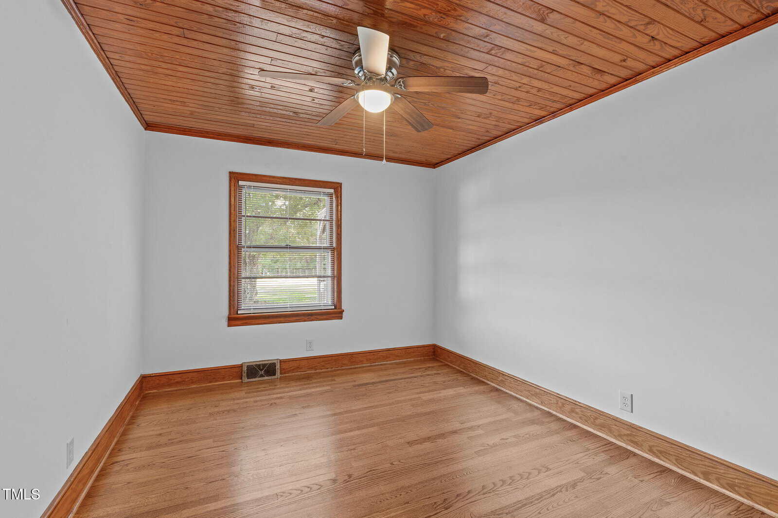 Empty room with light hardwood floors, wood-paneled ceiling, ceiling fan with light, white walls, baseboard trim, and a single window with a view of trees outside.