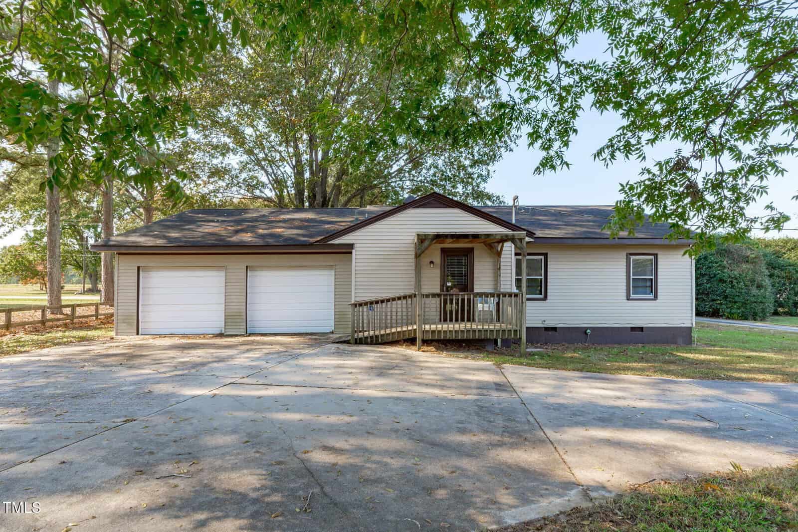 Single-story tan house with a small covered front porch, two-car garage, and paved driveway. Large trees and grassy areas surround the home. The weather appears clear and sunny.