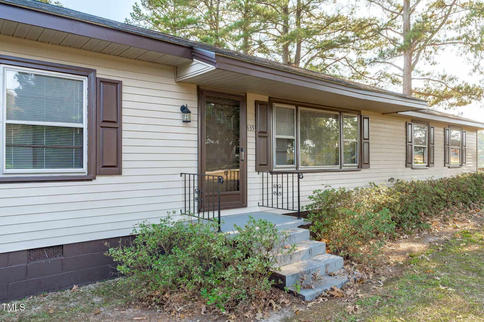 A single-story beige house with brown shutters and trim, featuring a front entrance with steps, a glass door, metal railings, and several large windows. Bushes line the front, and tall pine trees are visible in the background.