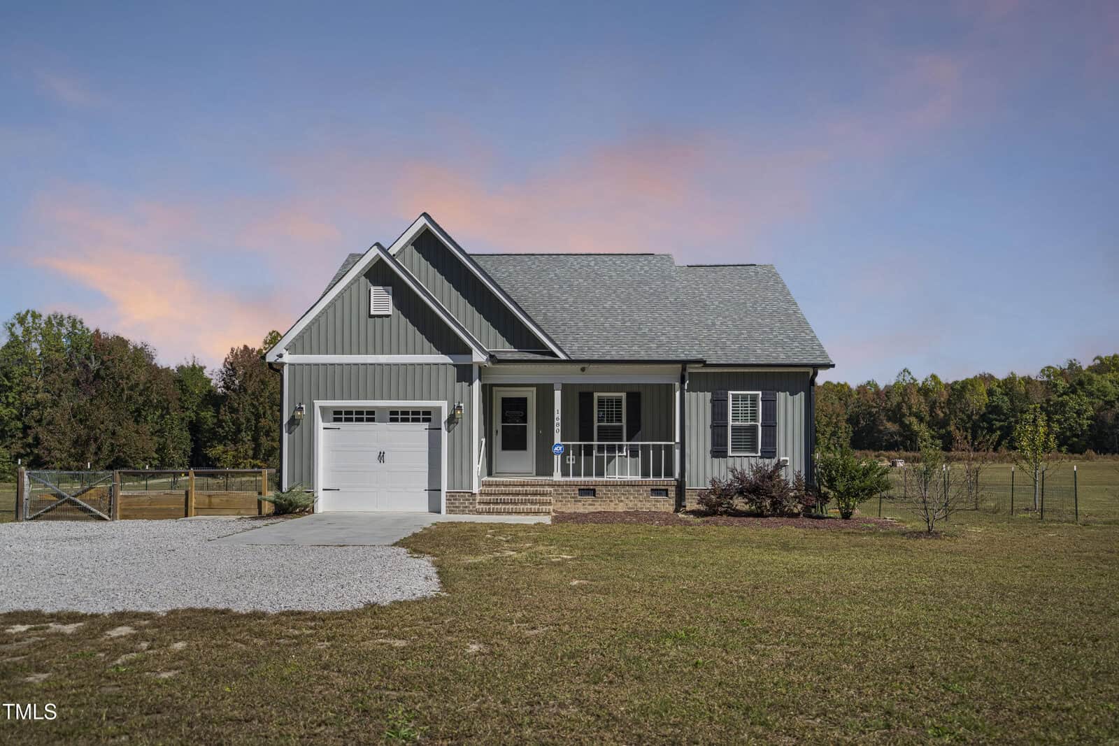 A small, single-story house with gray siding and a front porch. It has an attached garage and is surrounded by a large grassy yard. The sky is clear with a pinkish hue at the horizon, suggesting sunset or sunrise. Trees line the background.