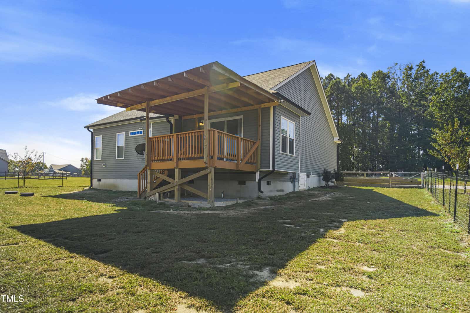 A house with a raised wooden deck and covered porch stands in a spacious grassy yard. The structure is a single-story with gray siding and white trim, surrounded by trees and neighboring houses under a clear blue sky.
