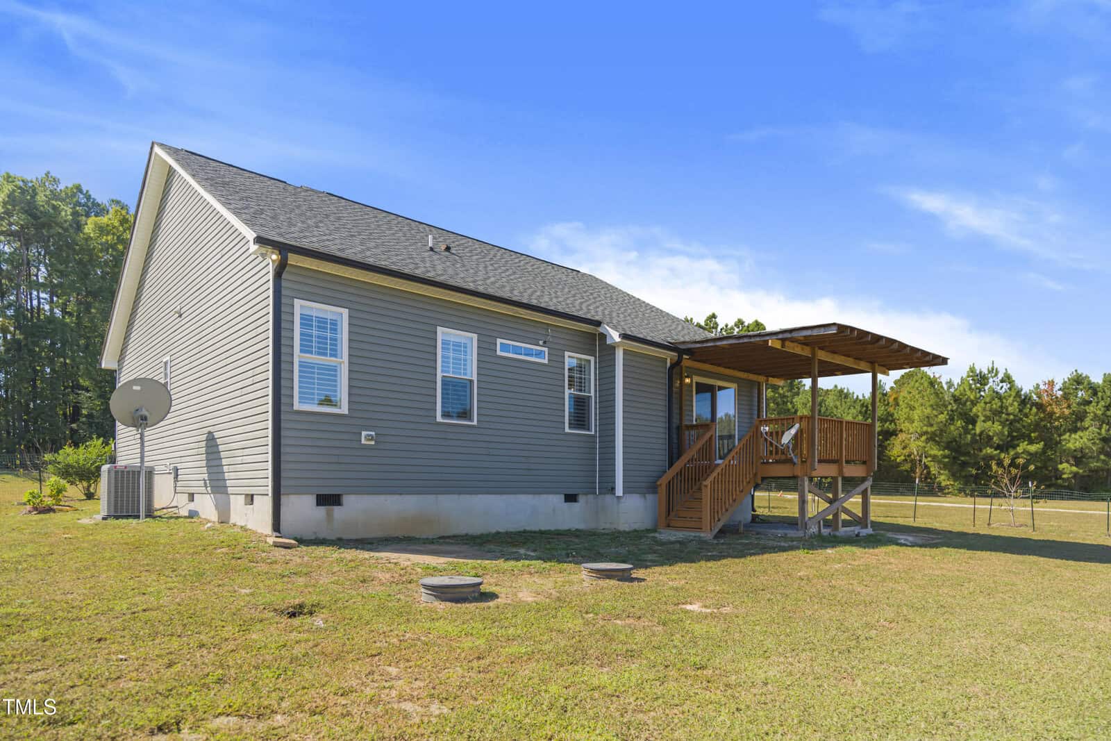 A single-story gray house with a sloped roof and a covered wooden deck in the backyard. There's a satellite dish on the side, and the house is surrounded by a grassy lawn and trees in the background under a clear blue sky.