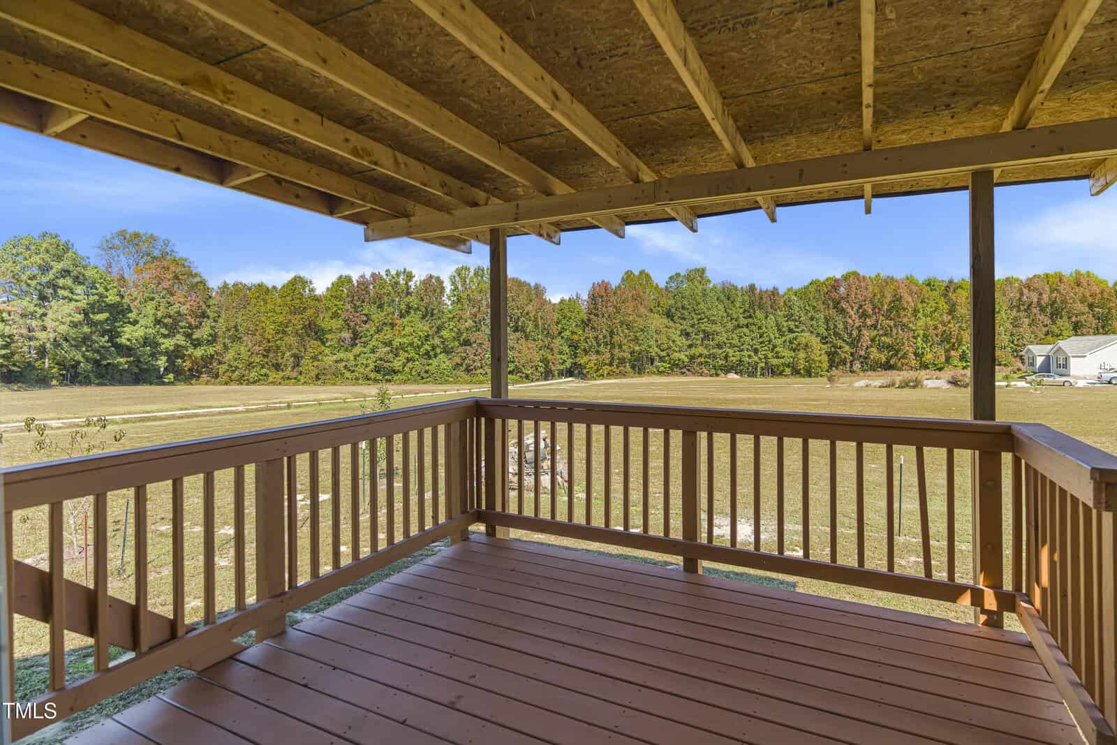 A wooden deck under a partially constructed roof overlooks an expansive grassy area. Trees with autumn foliage are visible in the distance under a clear blue sky.