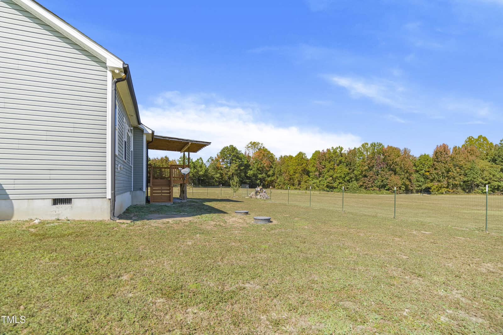 A side view of a house with beige siding and a covered porch. The house is next to a large, fenced grassy yard under a clear blue sky. Trees with autumn foliage line the background.