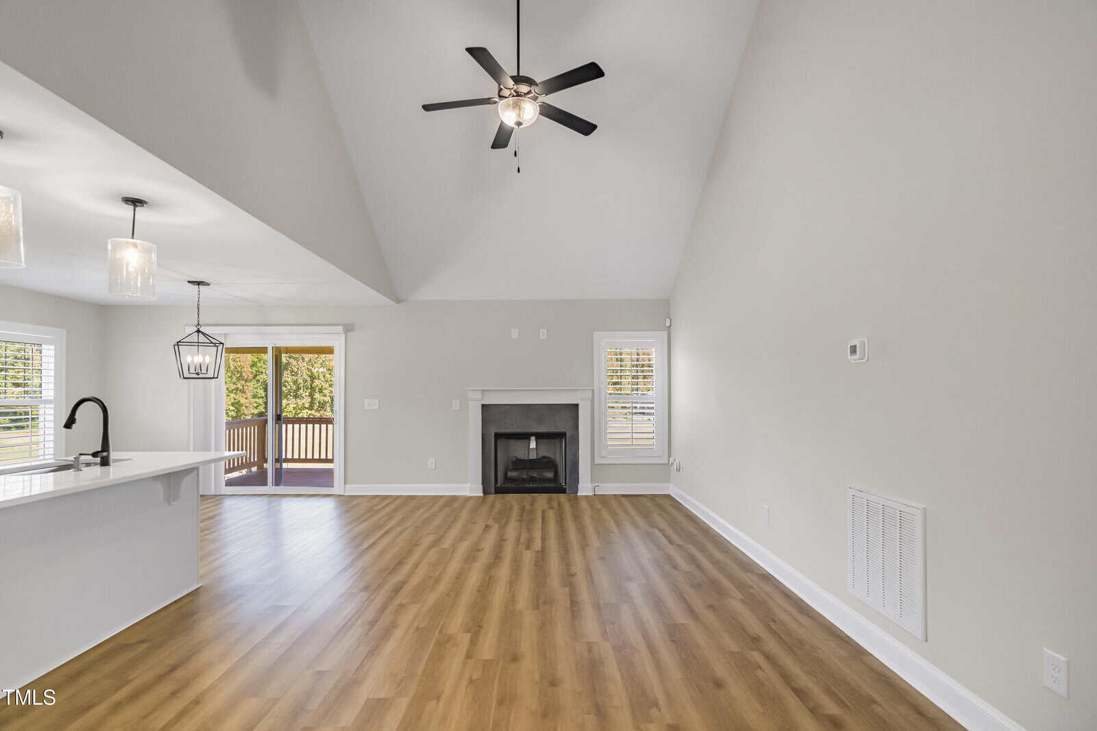 Spacious living room with vaulted ceiling, ceiling fan, and wood flooring. Includes a fireplace, sliding glass doors leading to a deck, and an open view of a kitchen with white cabinets and pendant lights over a marble countertop. Natural light fills the room.
