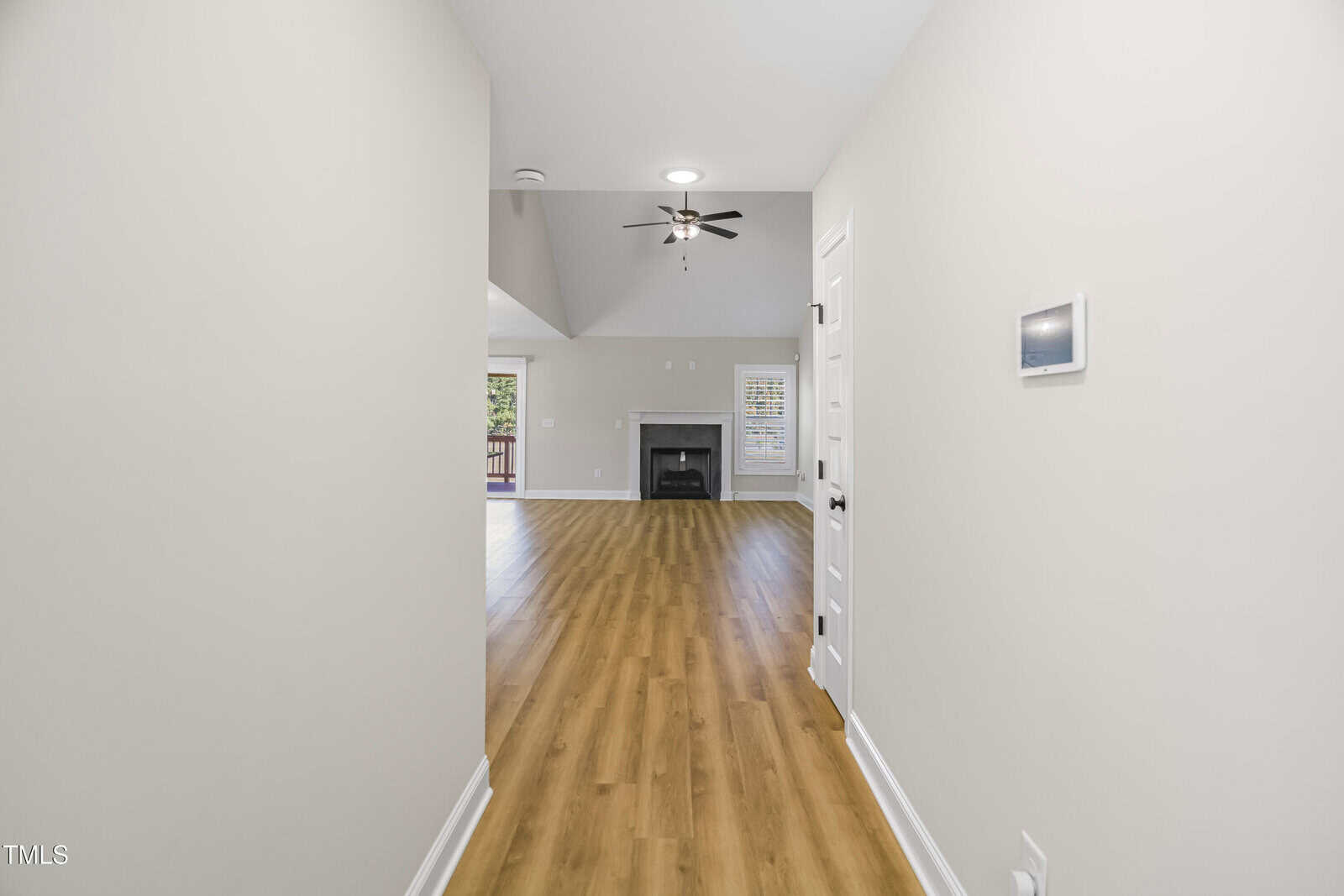 View of a long hallway with light wood flooring leading to an open living area. The room features a ceiling fan, a fireplace, large windows, and a door leading outside, creating a bright and airy atmosphere. Walls are painted a neutral color.