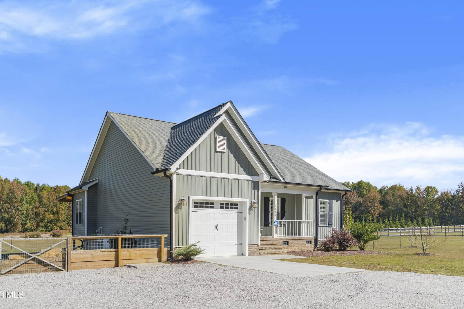 A small single-story house with gray siding and a gabled roof. It has a white front porch, a garage, and a gravel driveway. The house is surrounded by a grassy yard and a wooden fence, with trees and a blue sky in the background.
