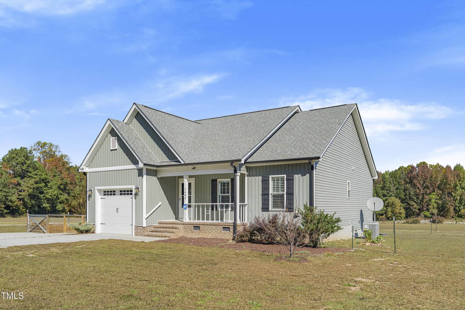 A single-story, light gray house with a gabled roof, white trim, and a garage. It has a small porch and is surrounded by a grassy yard. The sky is clear and blue.