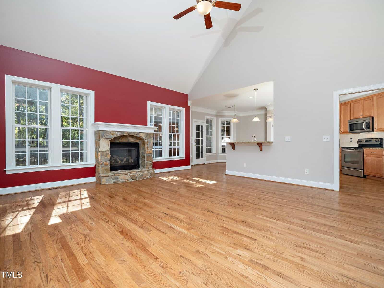 Spacious living room with hardwood floors, a red accent wall, and a stone fireplace. Features large windows and a ceiling fan. The open layout connects to a kitchen with stainless steel appliances and a breakfast bar.