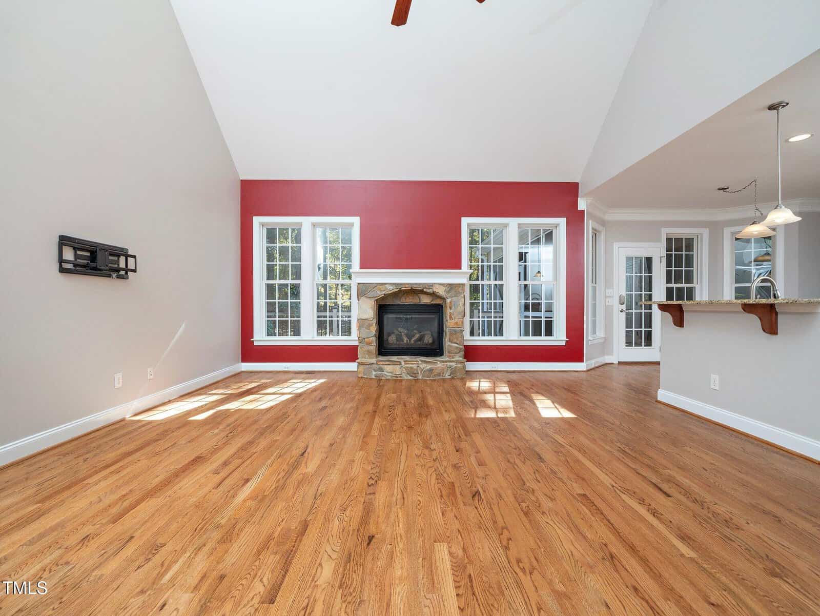 A spacious living room with hardwood floors, a red accent wall, and a stone fireplace. Large windows allow natural light to fill the room. The ceiling is vaulted, and pendant lights hang over a kitchen counter visible on the right.
