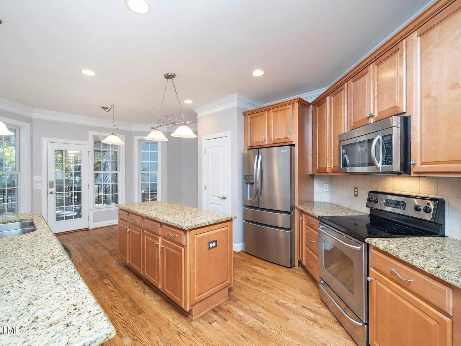 Spacious kitchen with granite countertops, wooden cabinets, stainless steel appliances, and a central island. French doors and multiple windows let in natural light. Hardwood floors and recessed lighting add to the modern design.