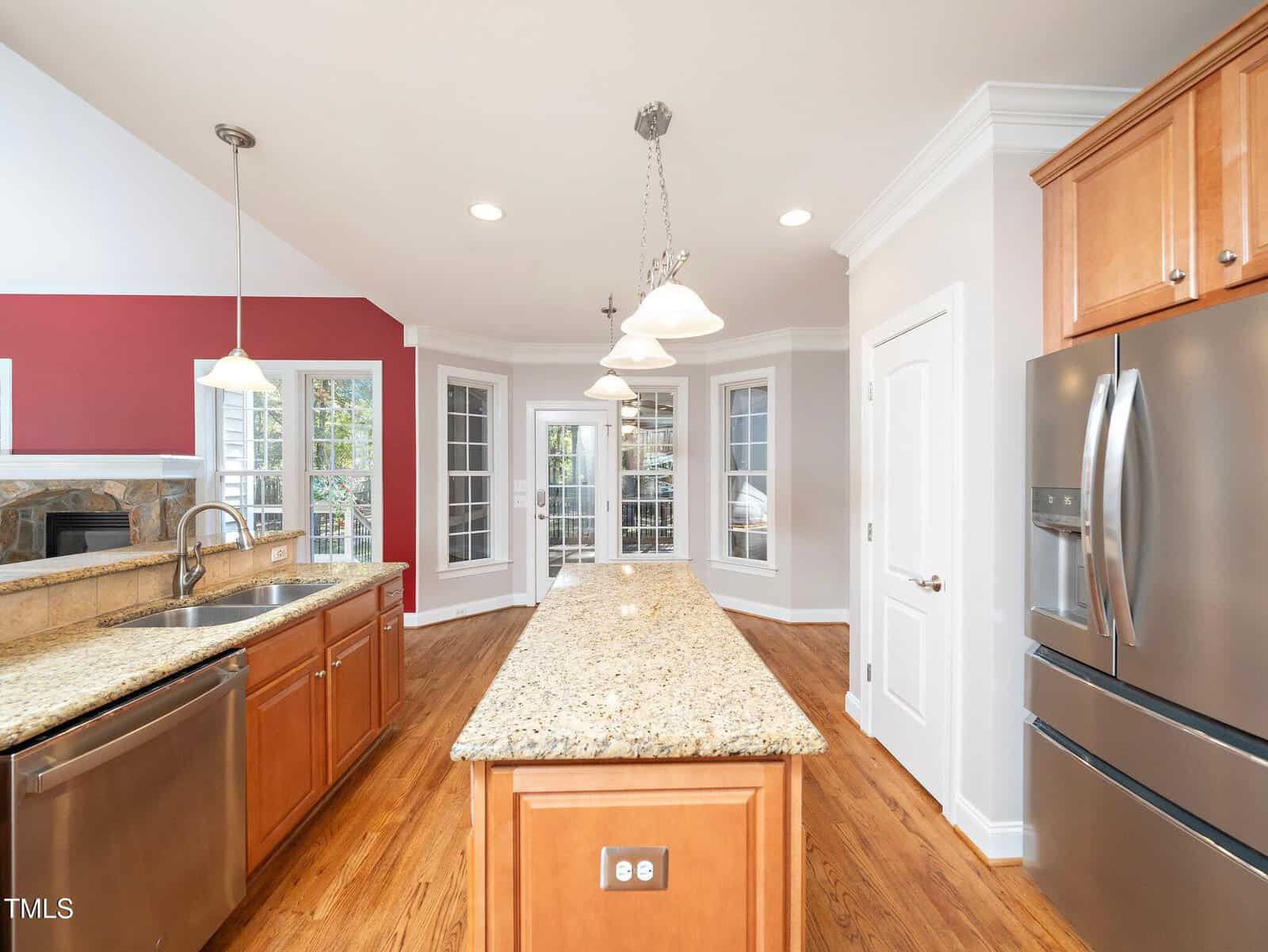 A spacious kitchen featuring a long granite island, stainless steel appliances, wooden cabinets, and hardwood floors. There's a red accent wall with large windows and French doors leading to a bright room. Pendant lights hang from the ceiling.