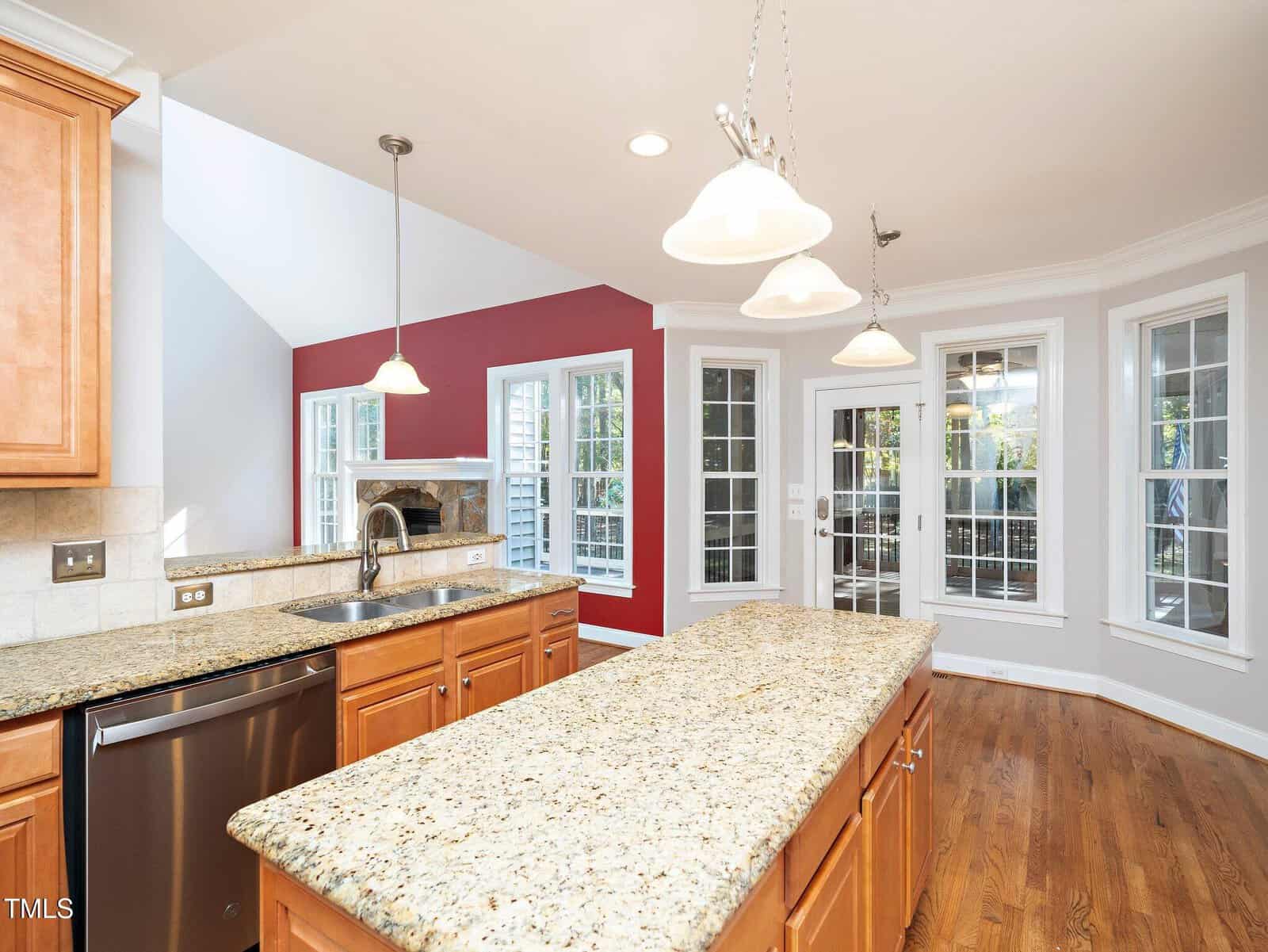A modern kitchen with a long granite island, wooden cabinets, and hardwood floors. Pendant lights hang above the island. Large windows and a red accent wall bring in natural light. A fireplace is visible in the background.