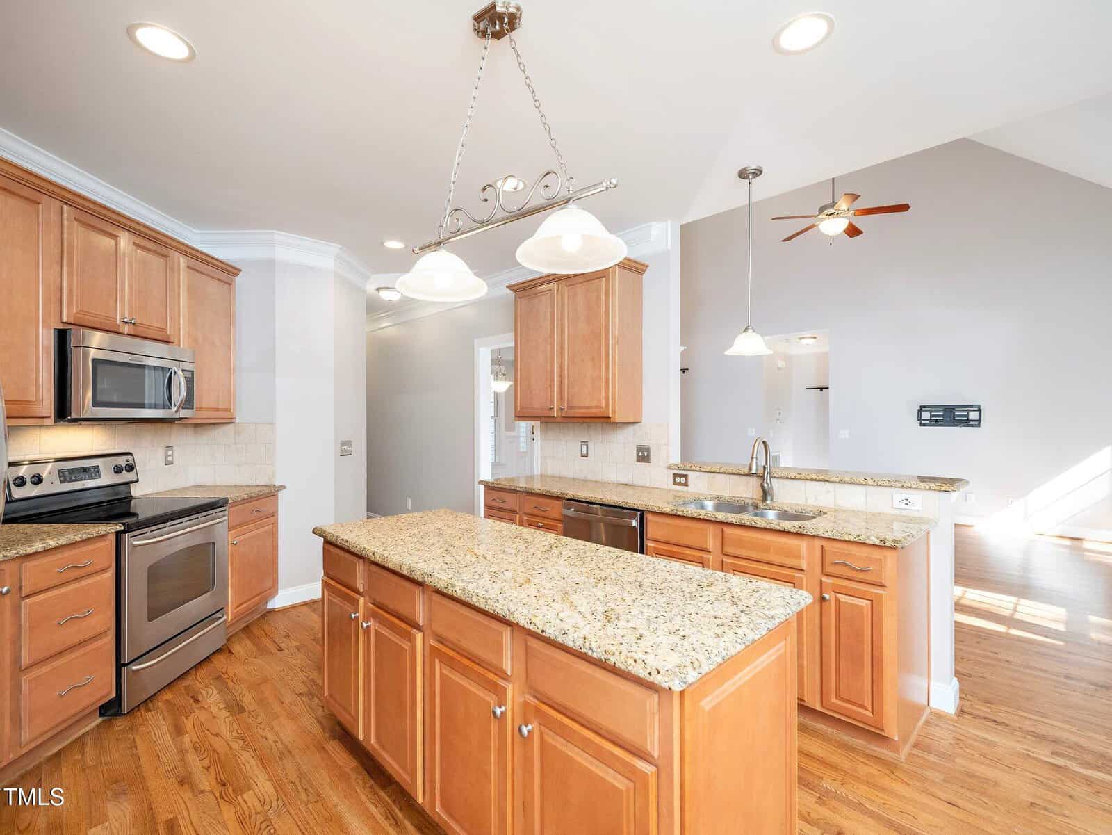 A spacious kitchen featuring wooden cabinets, a large granite-topped island, stainless steel appliances, and wooden flooring. Overhead lighting illuminates the area, and there is an open view into a brightly lit living space with a ceiling fan.