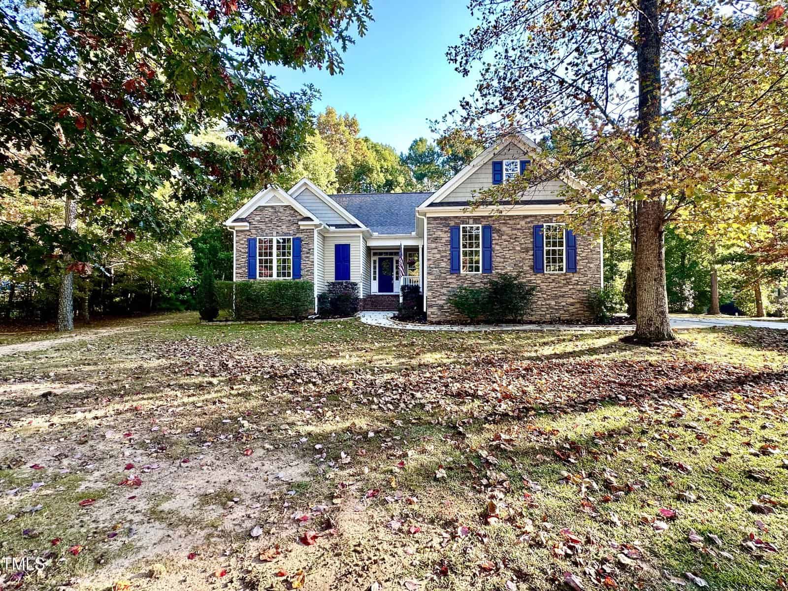 A charming single-story house with a brick facade, blue shutters, and a gray roof, set amidst a yard covered in fallen leaves. Surrounded by trees, the home exudes a cozy and inviting ambiance, enhanced by the bright afternoon sunlight.