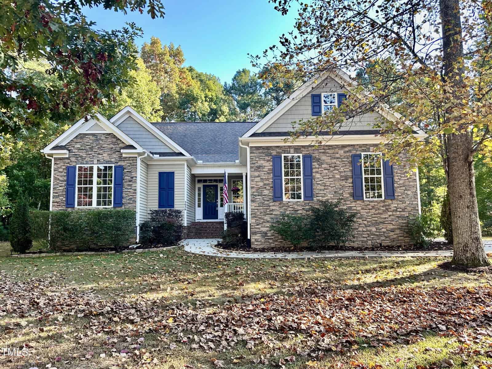 Nestled at 3636 Armida Drive in Wake Forest, this charming single-story house boasts a mix of stone and siding exterior. It features blue shutters and a covered front porch, all surrounded by trees and fallen leaves on the lawn, beautifully capturing the essence of autumn.