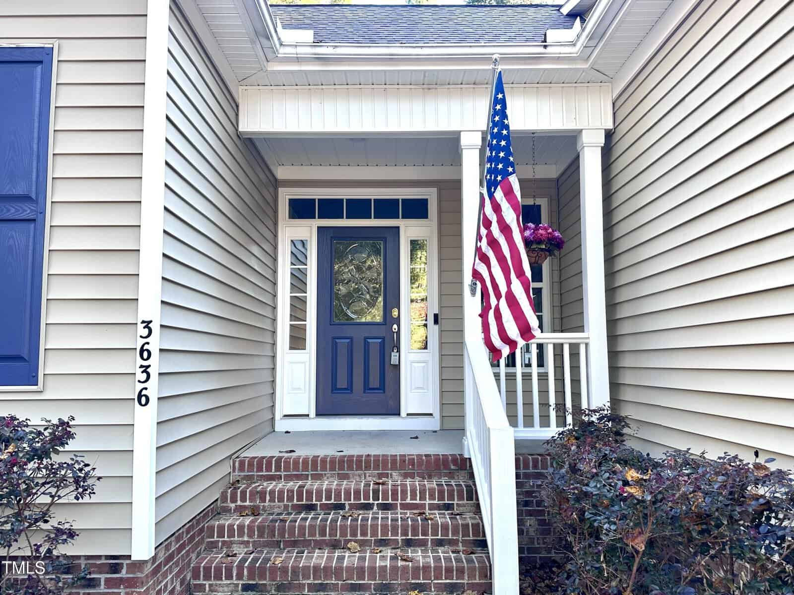 A house entrance with a dark blue door and a decorative glass panel above it. An American flag hangs beside the steps leading to the door. The exterior is beige siding with brick steps, and there are plants and a hanging basket with flowers.