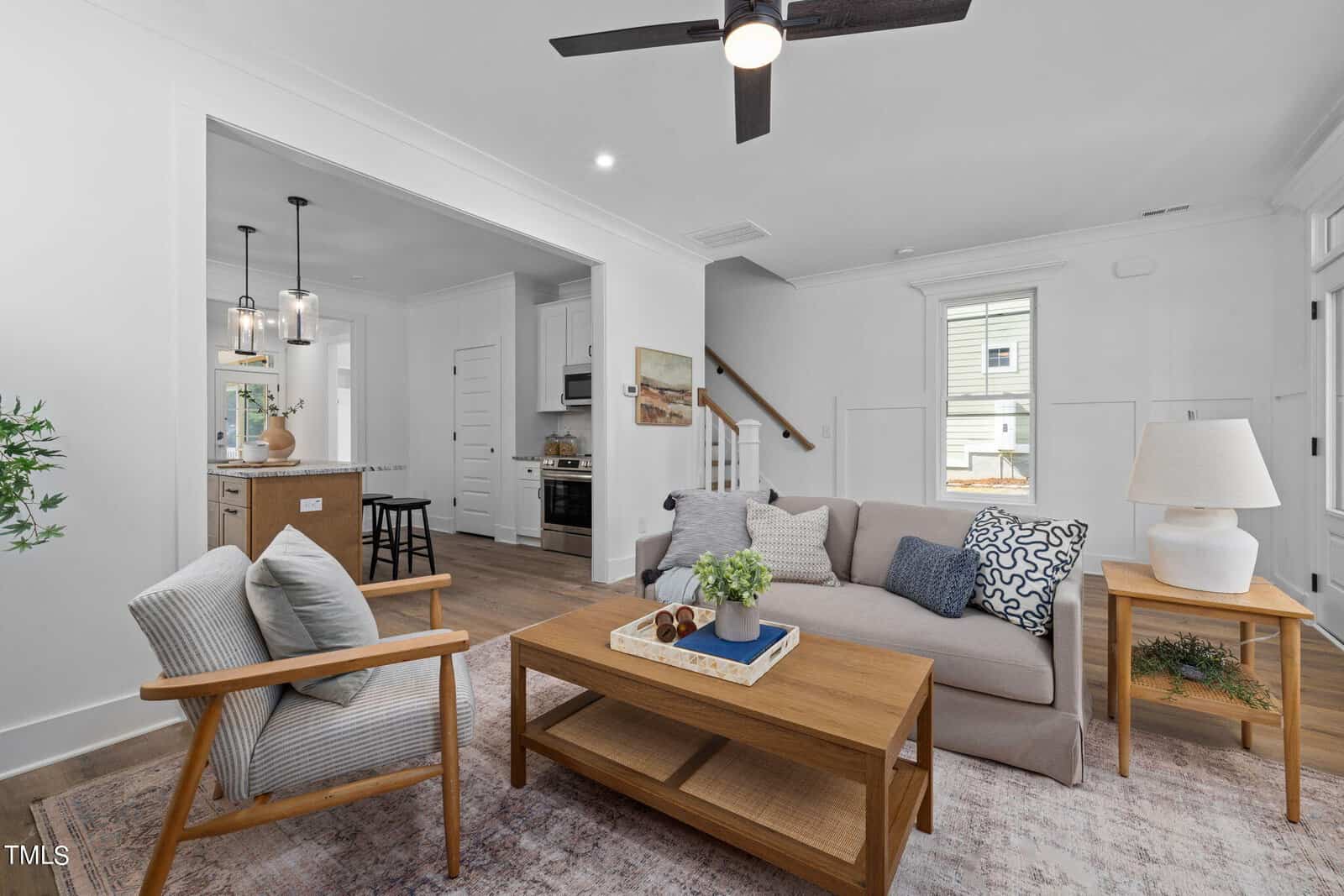 Modern living room with neutral tones, featuring a sofa, wooden coffee table, armchair, and side tables. Kitchen with island and pendant lights visible in the background; ceiling fan overhead and stairs to the side.