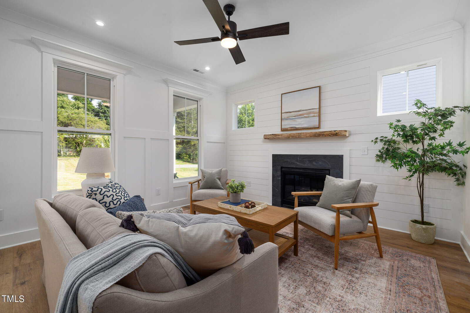 Bright, modern living room with white walls, shiplap accents, a gray sofa, two wooden armchairs, a coffee table, potted plant, fireplace with wooden mantel, large windows, and ceiling fan.