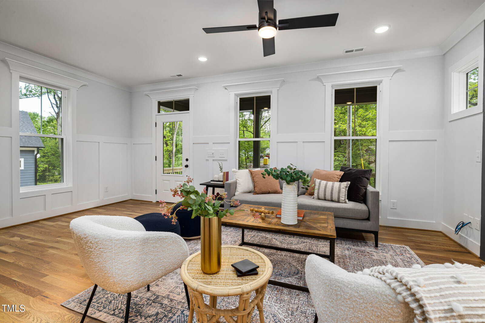 Modern living room with white walls, wood floors, and large windows. Features a gray sofa with pillows, two round chairs, a gold side table with a plant, and a textured rug. Ceiling fan and greenery visible outside.