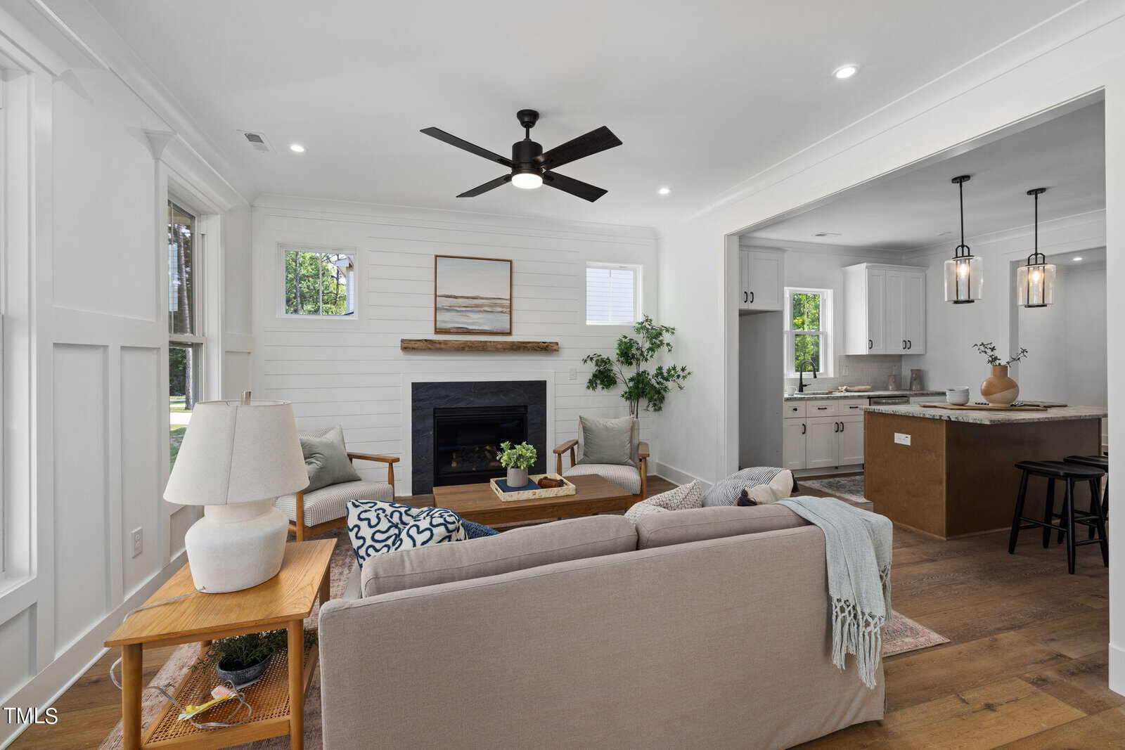 A bright, modern living room with light walls, wood floors, a beige sofa, and a fireplace. Large windows let in natural light. An open doorway leads to a contemporary kitchen with an island and pendant lights.