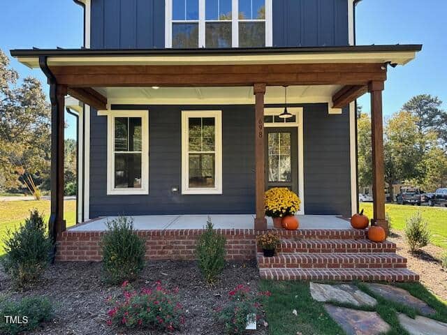 A modern house with dark siding and a wooden porch, decorated with orange pumpkins and yellow flowers. Brick steps lead to the front door, and the yard has bushes, small plants, and a stone path.