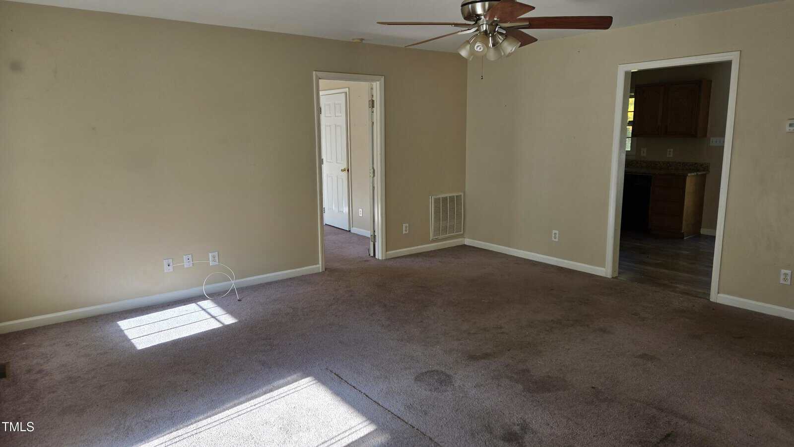 A vacant living room with beige walls and a ceiling fan. The carpet is brown and has noticeable stains. Sunlight enters through a window on the left. An open door leads to another room, and a partially visible kitchen is to the right.
