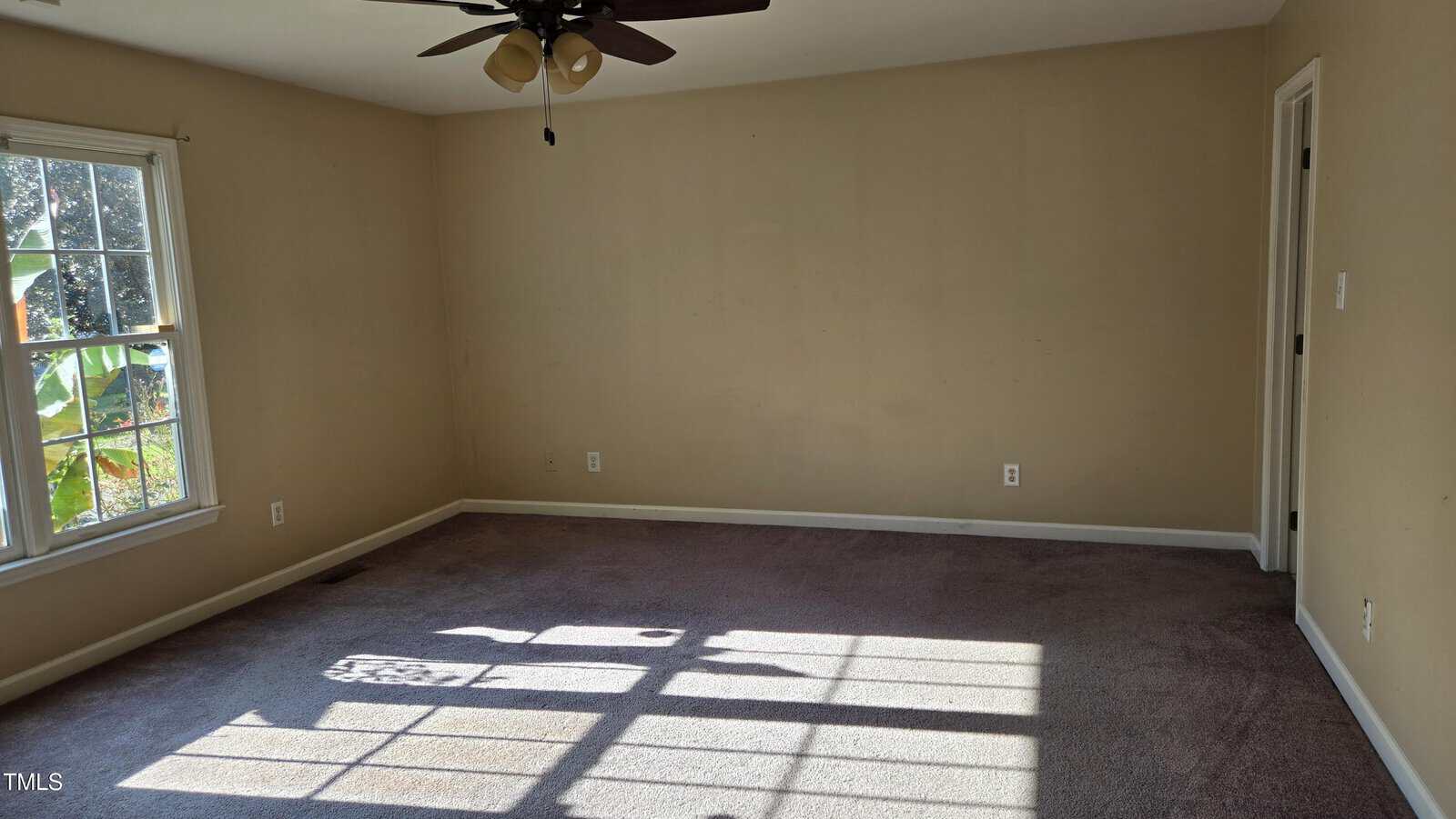 An empty room with beige walls and carpeted flooring. A ceiling fan with lights is centered on the ceiling. A large window on the left lets in sunlight, casting shadows on the floor, while a door is slightly ajar on the right.
