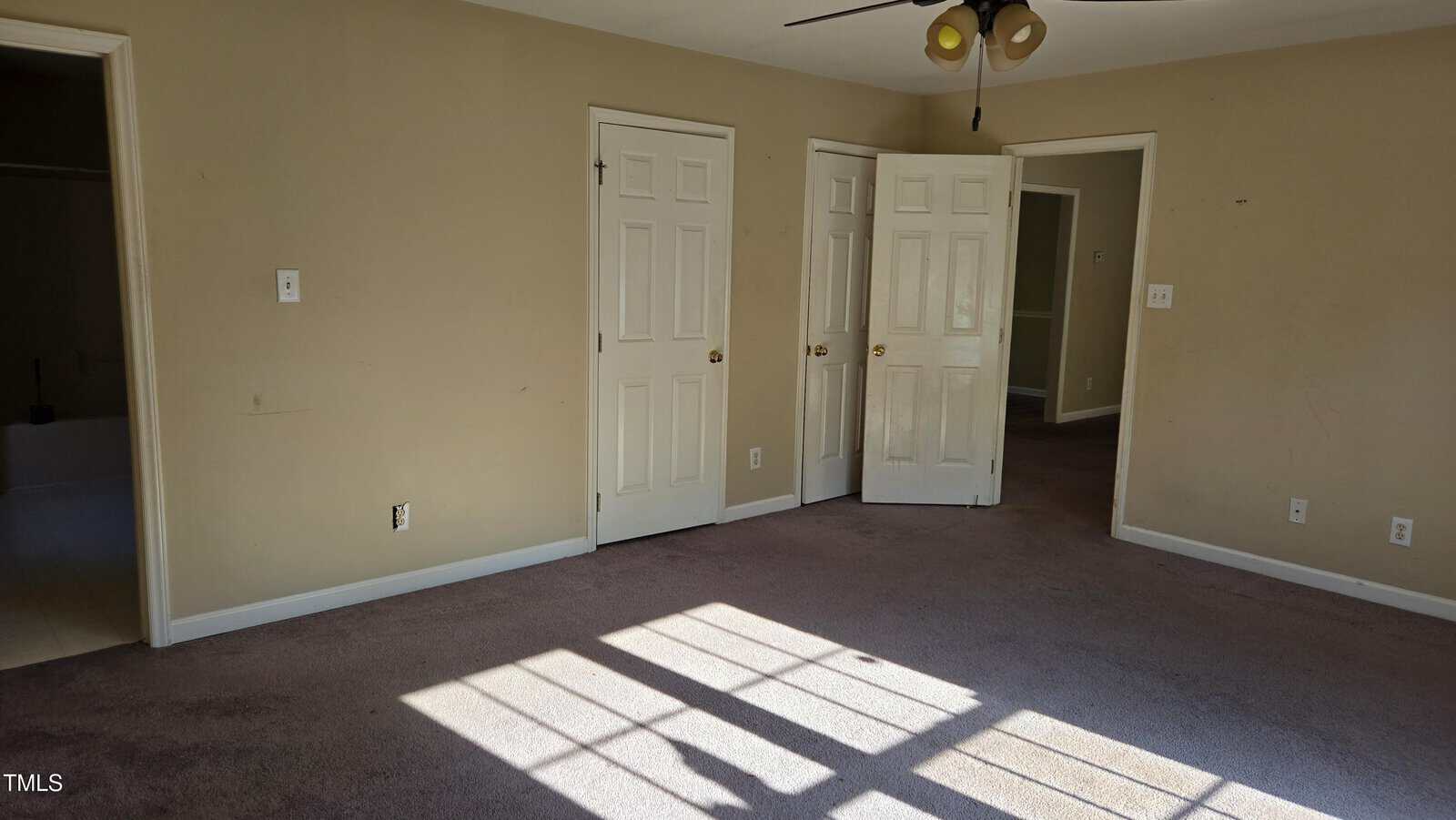 An empty room with beige walls and carpeting. The space features two closed doors and one open door leading to another area. Sunlight streams in through a window, casting shadows on the floor. A ceiling fan is partially visible.