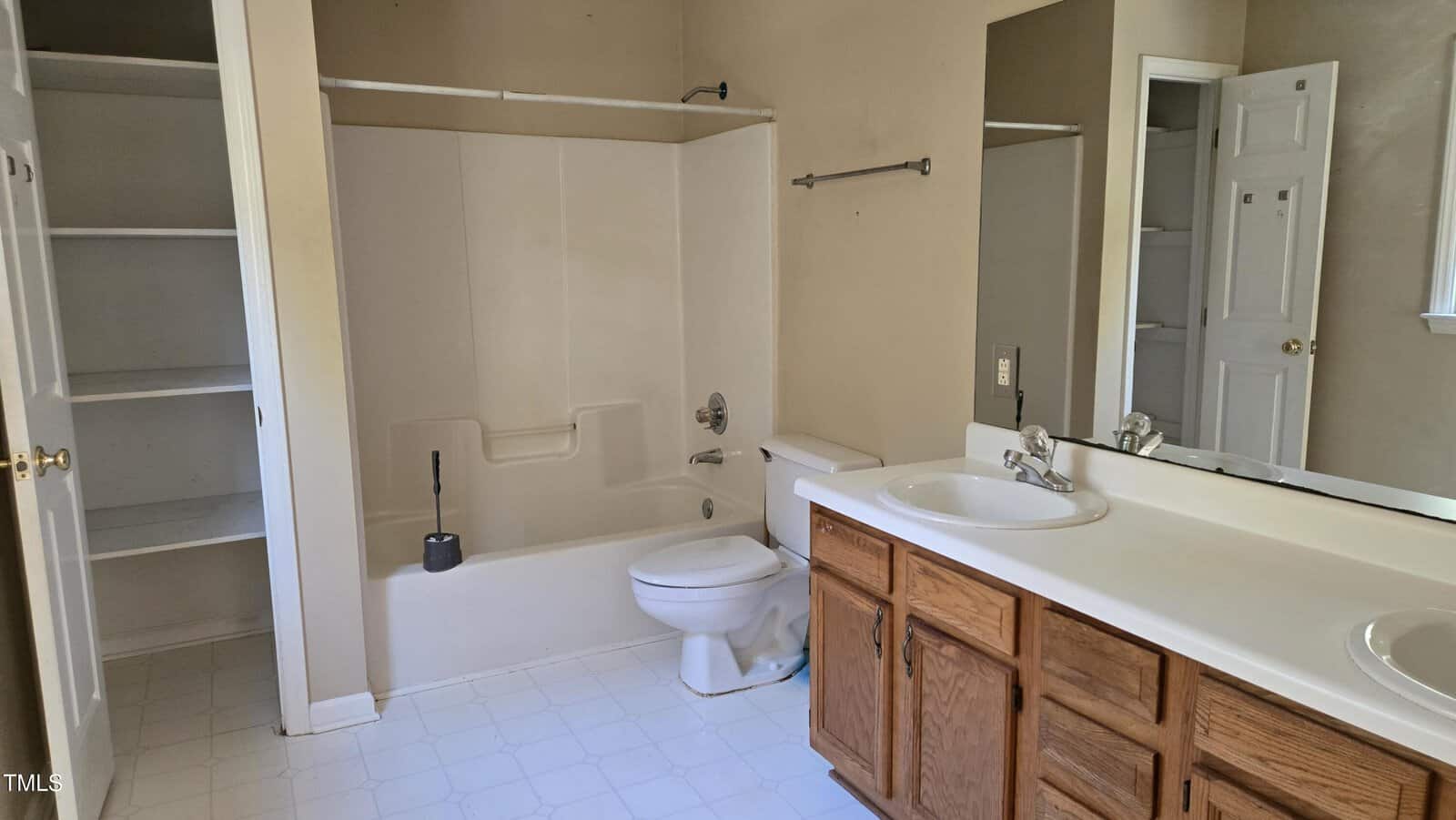 A bathroom with a bathtub-shower combination, toilet, and a double-sink vanity with wooden cabinets. The walls are beige, and the floor is white tile. There is a small closet with open shelving visible on the left.