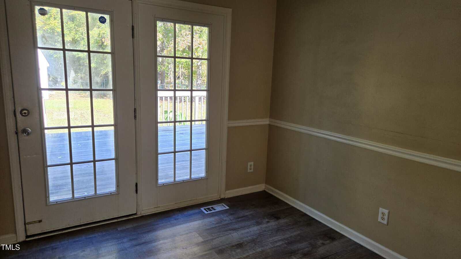A room with beige walls and white trim features double glass doors leading to a deck. The floor is covered with dark wood-style vinyl, and there is an electrical outlet and a small vent on the wall. Natural light comes through the doors.