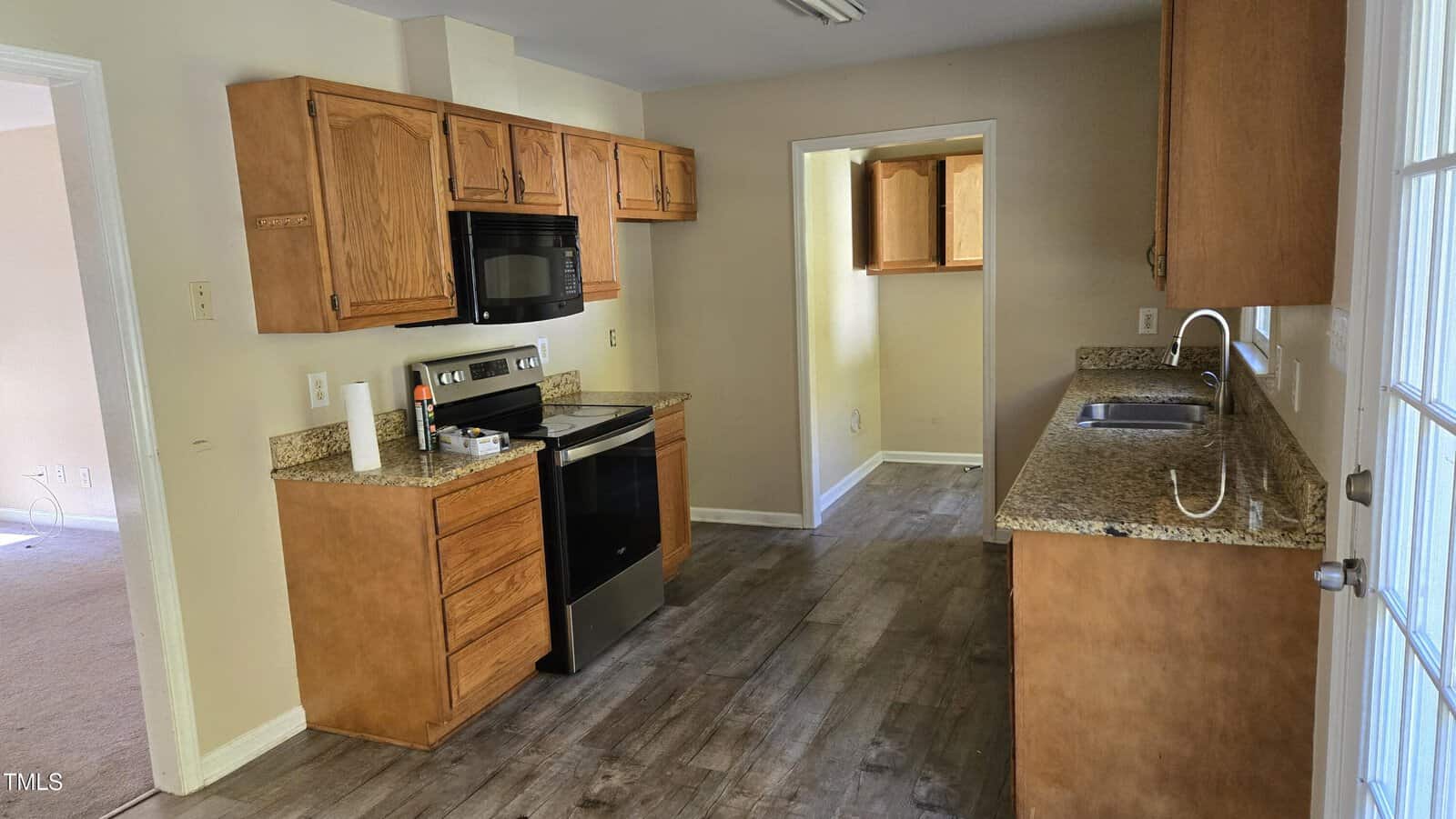 A kitchen with wooden cabinets and granite countertops. It has a stainless steel stove with a black microwave above. There is a sink on the right, and the floor is made of dark wood. A doorway leads to another room at the back.