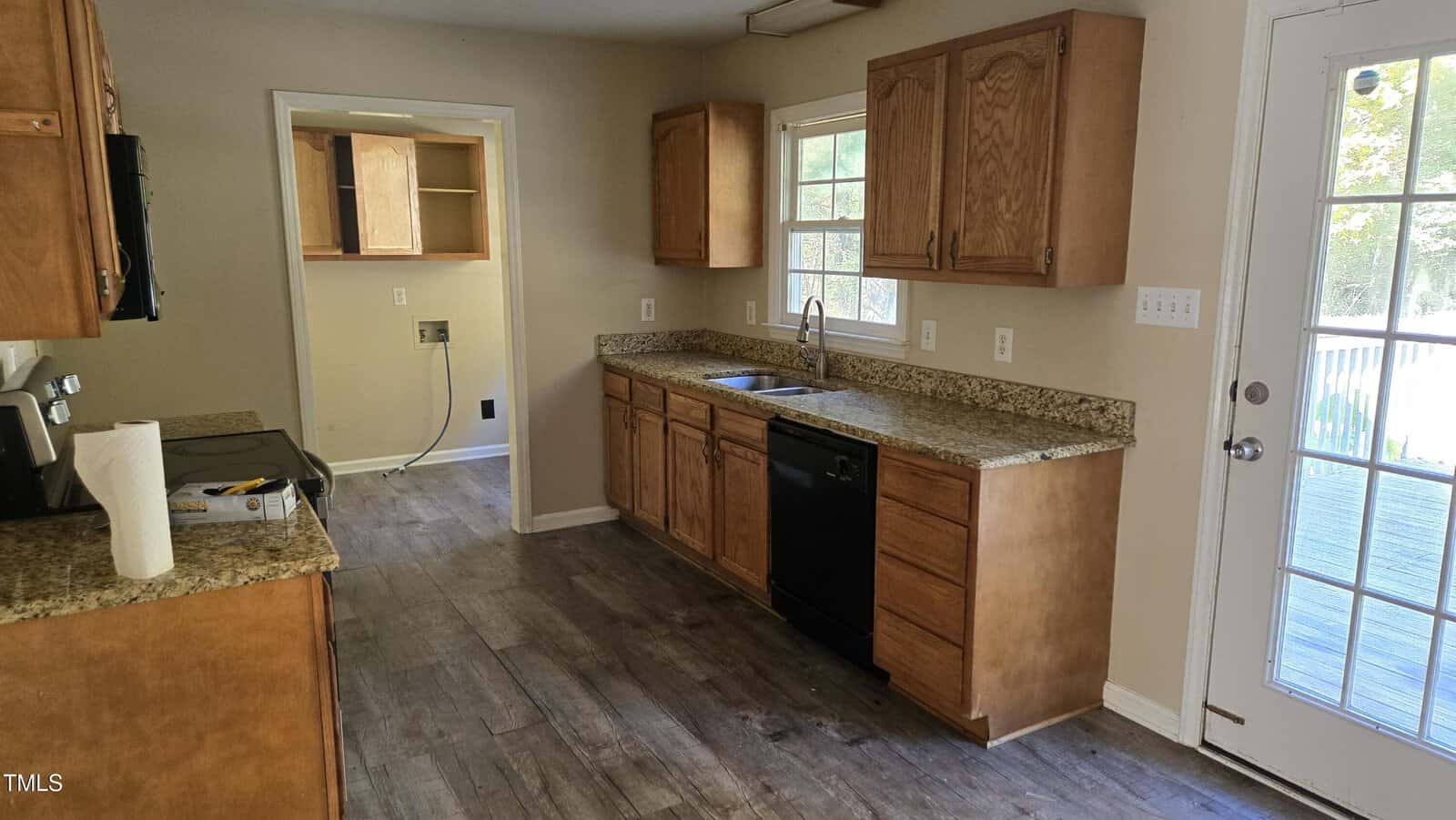 A kitchen featuring wooden cabinets, granite countertops, and a stainless steel oven. A black dishwasher is integrated below the counter near the sink. The kitchen leads to a door with glass panes that opens to the outside. The floor has a wood-like finish.