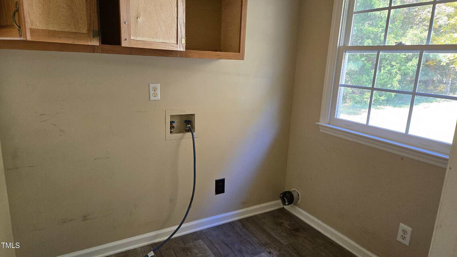 A vacant laundry area with a washing machine hookup and an empty cabinet above. A power outlet and plumbing connections are visible on the wall. The room has beige walls, and a window on the right side allows sunlight to illuminate the space.