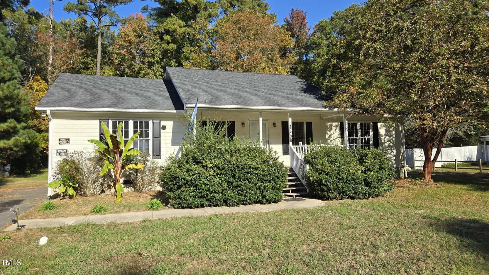 A single-story beige house with a dark gray roof is surrounded by green shrubs and trees. The front porch has white railings and steps. A driveway is visible on the left, and the background features tall trees with some autumn leaves.